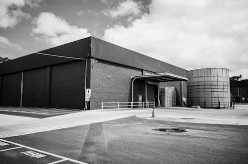 Free Black and white photo of a large industrial warehouse with a storage tank and parking area. Stock Photo