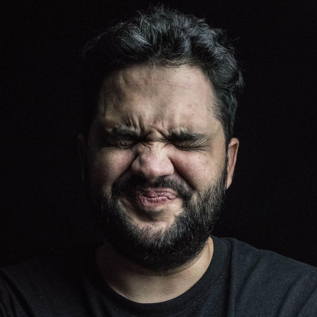 Close-up portrait of a man with facial hair showing a quirky expression in dim lighting.