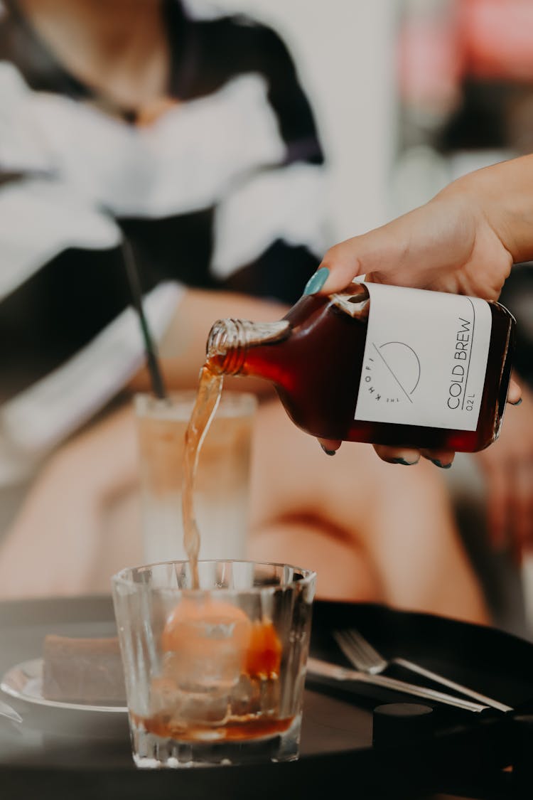 A Person Pouring Coffee From A Bottle To A Glass Cup With Ice