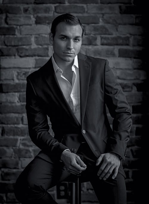 Grayscale Photo of a Man in a Suit Sitting Near a Brick Wall