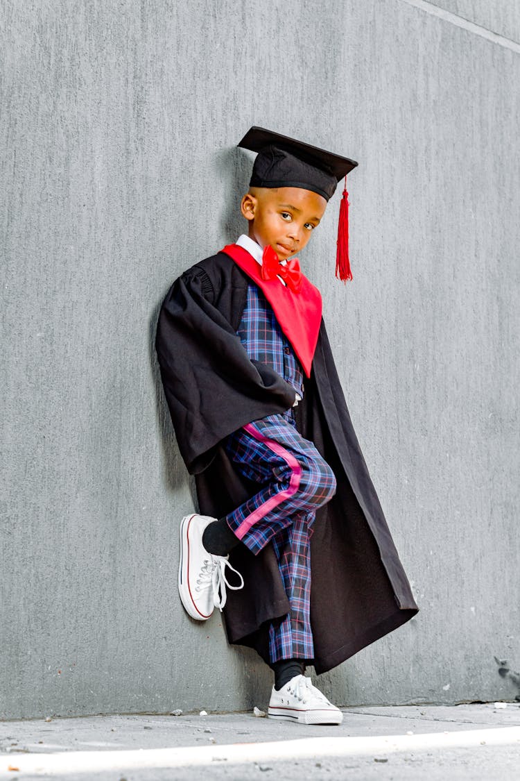 Boy In Black Graduation Gown And Square Academic Cap Leaning On Gray Wall