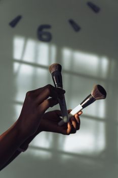 Close-up of hands holding makeup brushes against a soft-lit clock background.