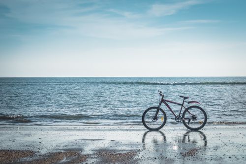 Free A lone bicycle rests on a serene beach with a calm ocean and blue sky. Stock Photo