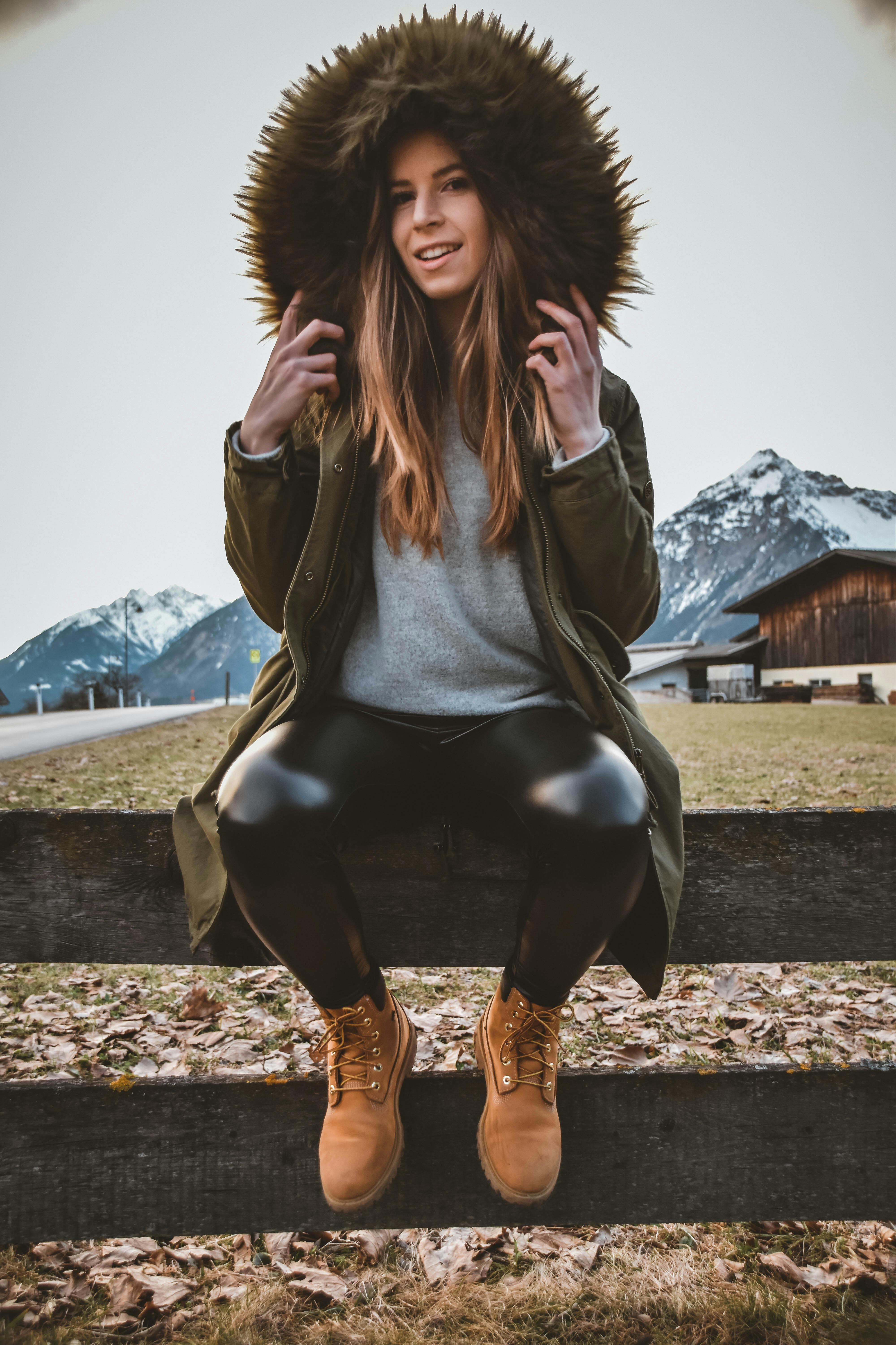 woman wearing brown and green parka sitting on gray wooden fence at daytime