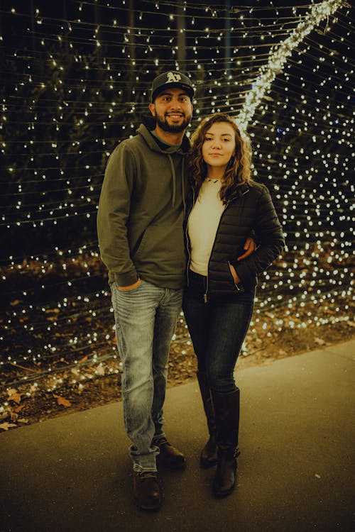 A Couple Standing Beside Each Other Near String Lights while Looking at the Camera