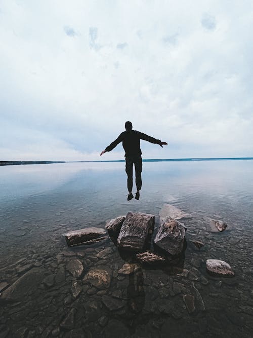 Man in Black Jacket and Pants Standing on Rock Near Body of Water