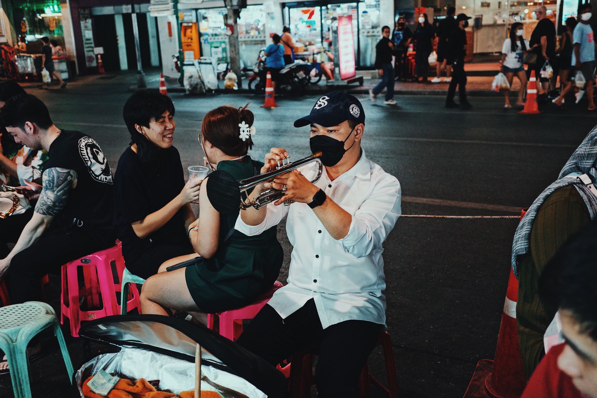A street musician wearing a face mask plays the trumpet in a lively night market scene.
