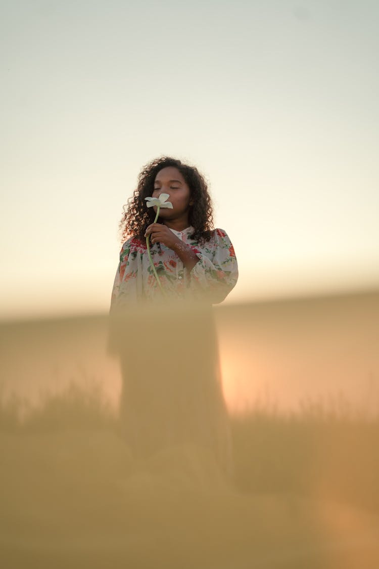 Woman Standing In Field And Smelling White Flower In Her Hand