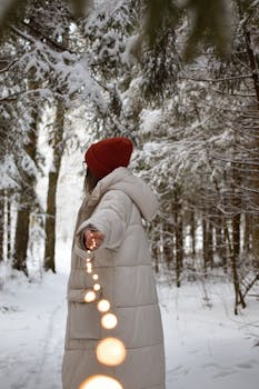 A woman in a winter coat and hat holds string lights in a snowy forest, creating a magical winter scene.
