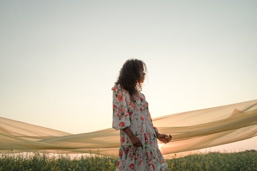 Woman in Floral Dress Walking in Flower Field with Tulle Fabric in Background