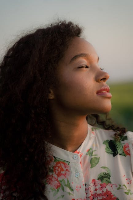 A pensive young woman with curly hair in floral attire enjoys a sunset outdoors.