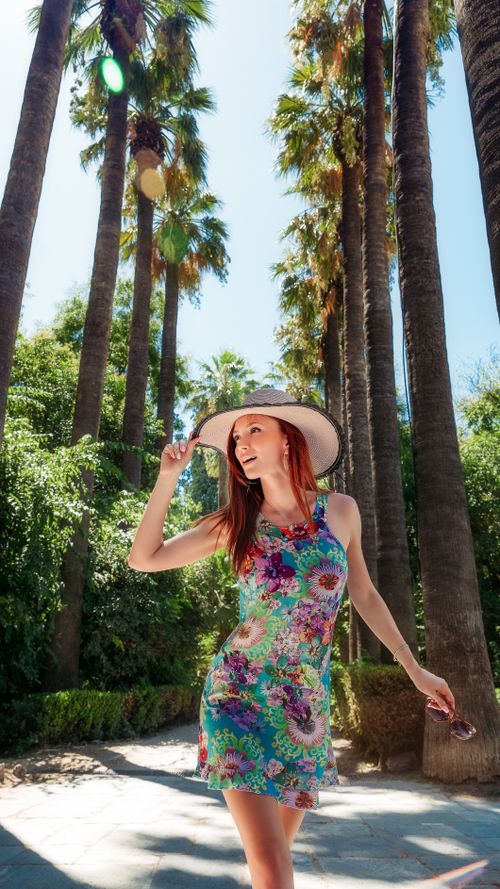 Free A woman in a floral dress and hat enjoying sunny weather in a palm-lined garden in Athens. Stock Photo
