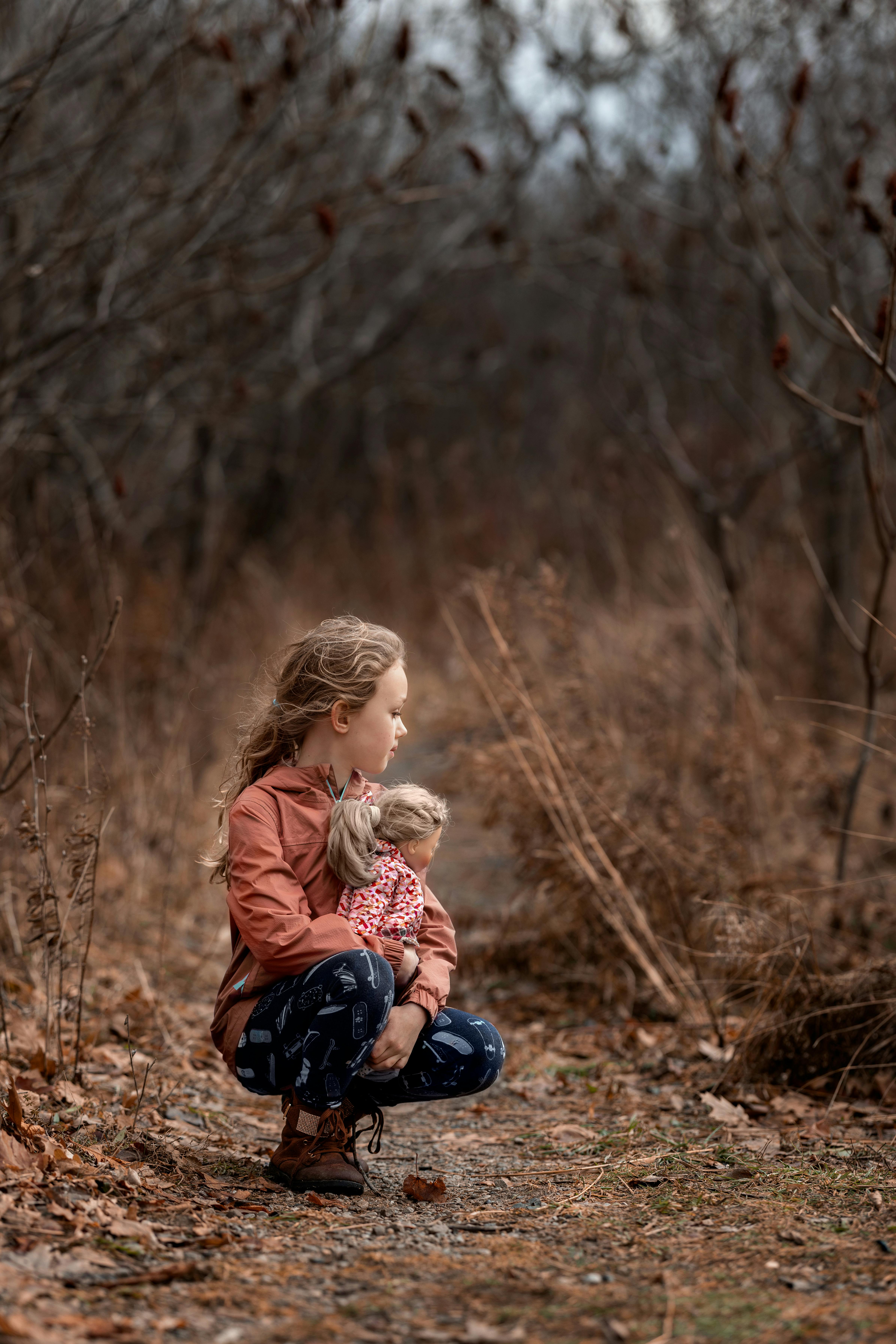 little girl holding a doll crouching on a path in forest