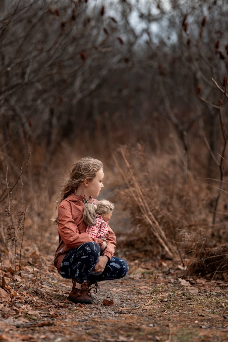 Little Girl Holding A Doll Crouching On A Path In Forest 