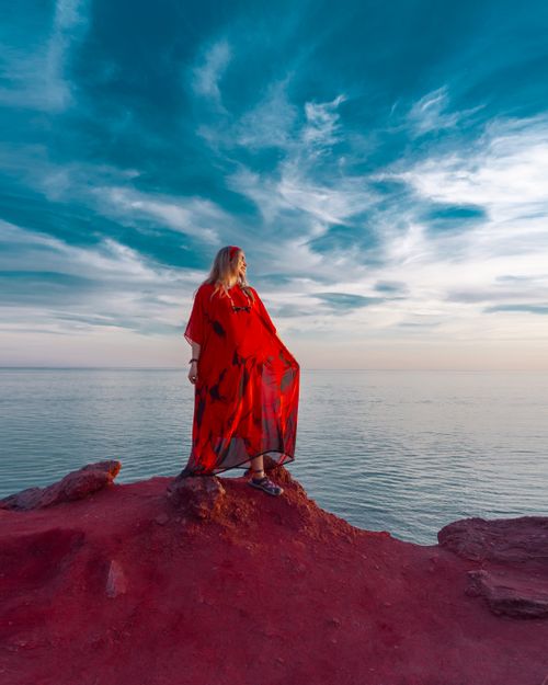 Free Serene view of a woman in a red dress overlooking the Persian Gulf during dusk with vibrant red cliffs. Stock Photo