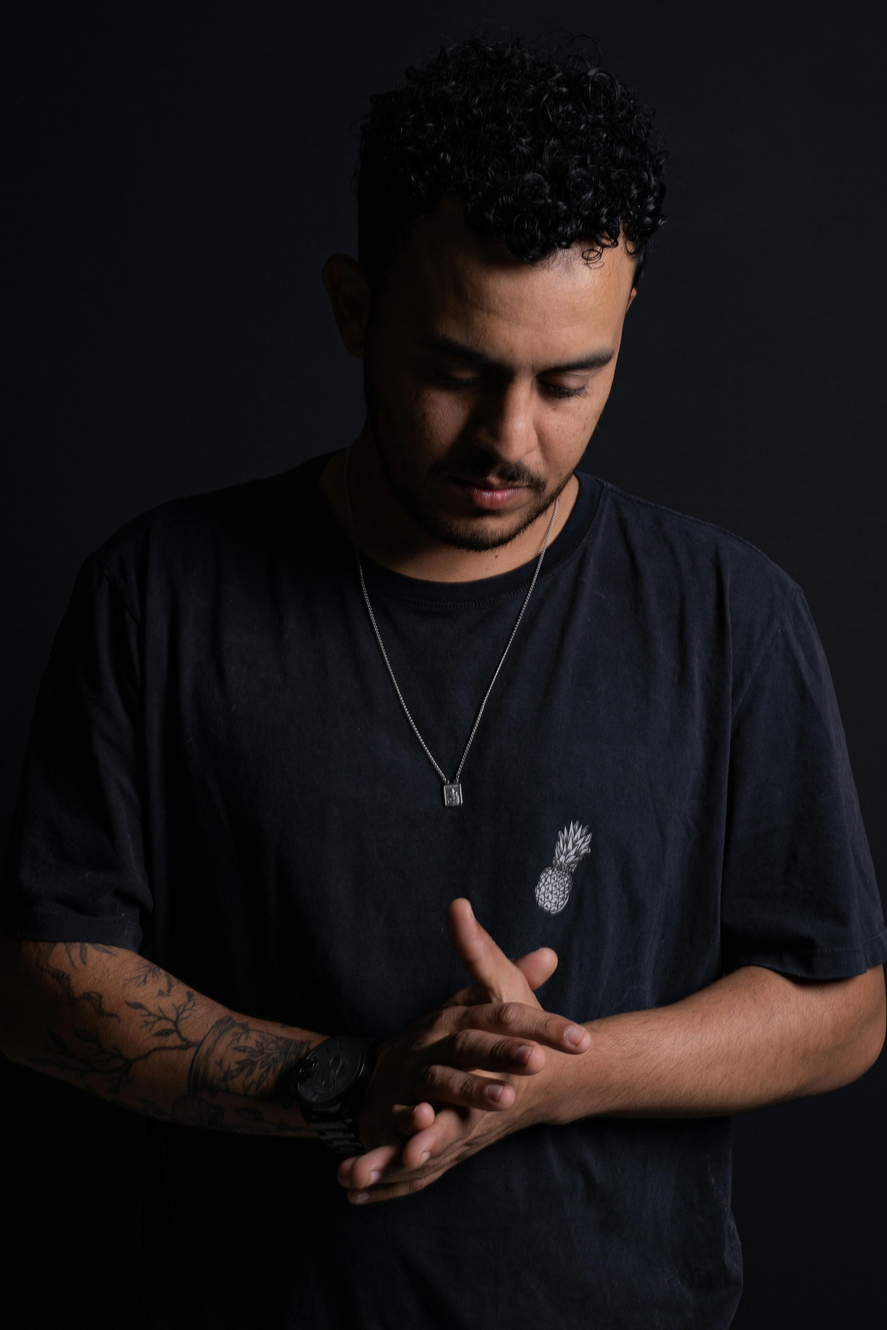 Striking portrait of a man standing indoors in a dark setting, showcasing his tattoos and casual style.