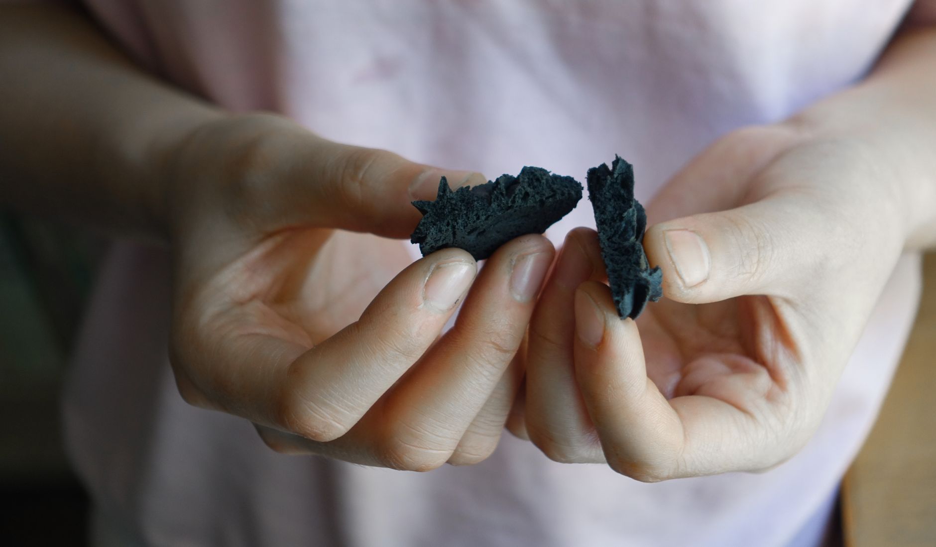 A close-up image showing hands holding a broken charcoal biscuit against a blurred background.