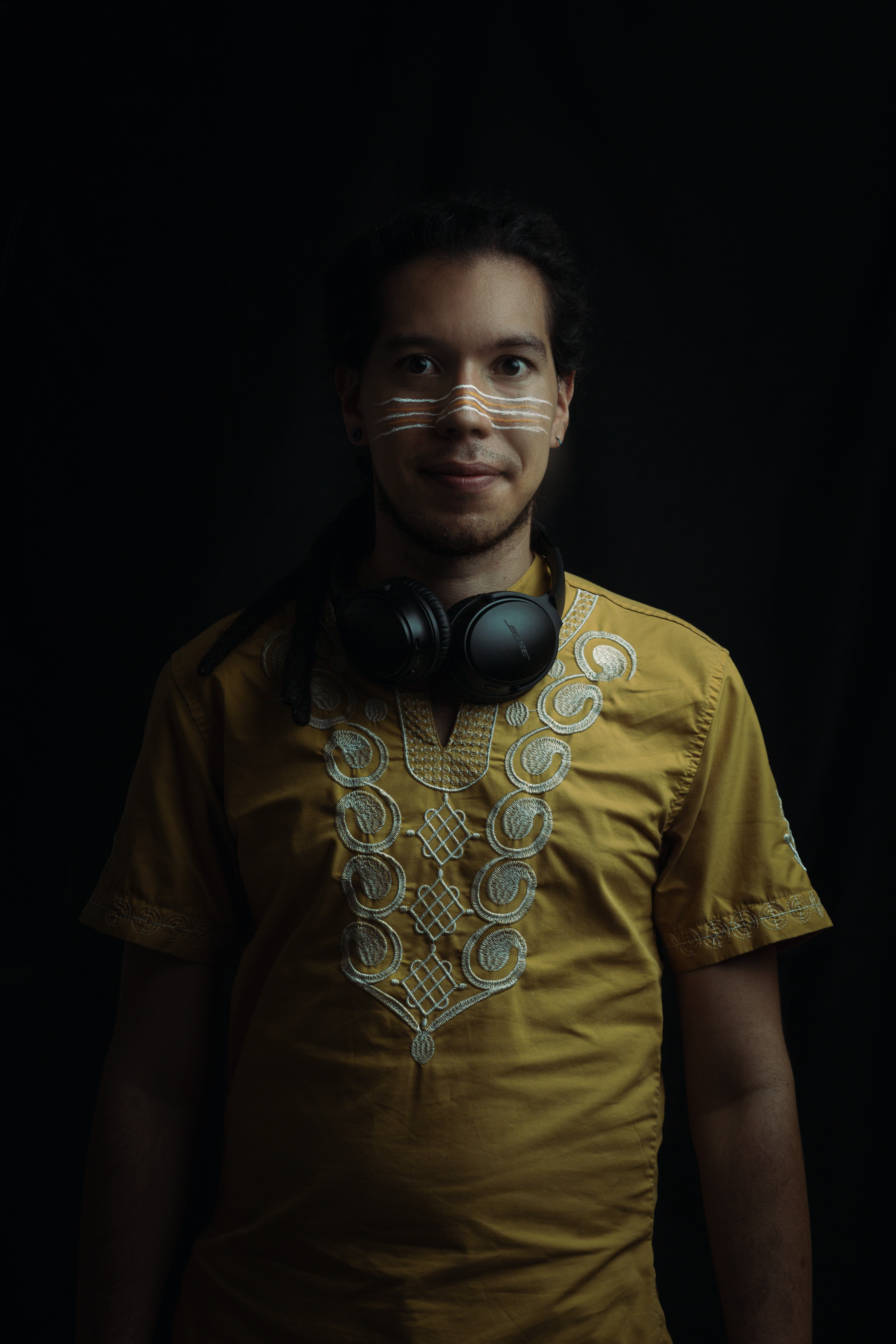 Studio portrait of a man wearing headphones around his neck and an ethnic shirt, set against a dark background.