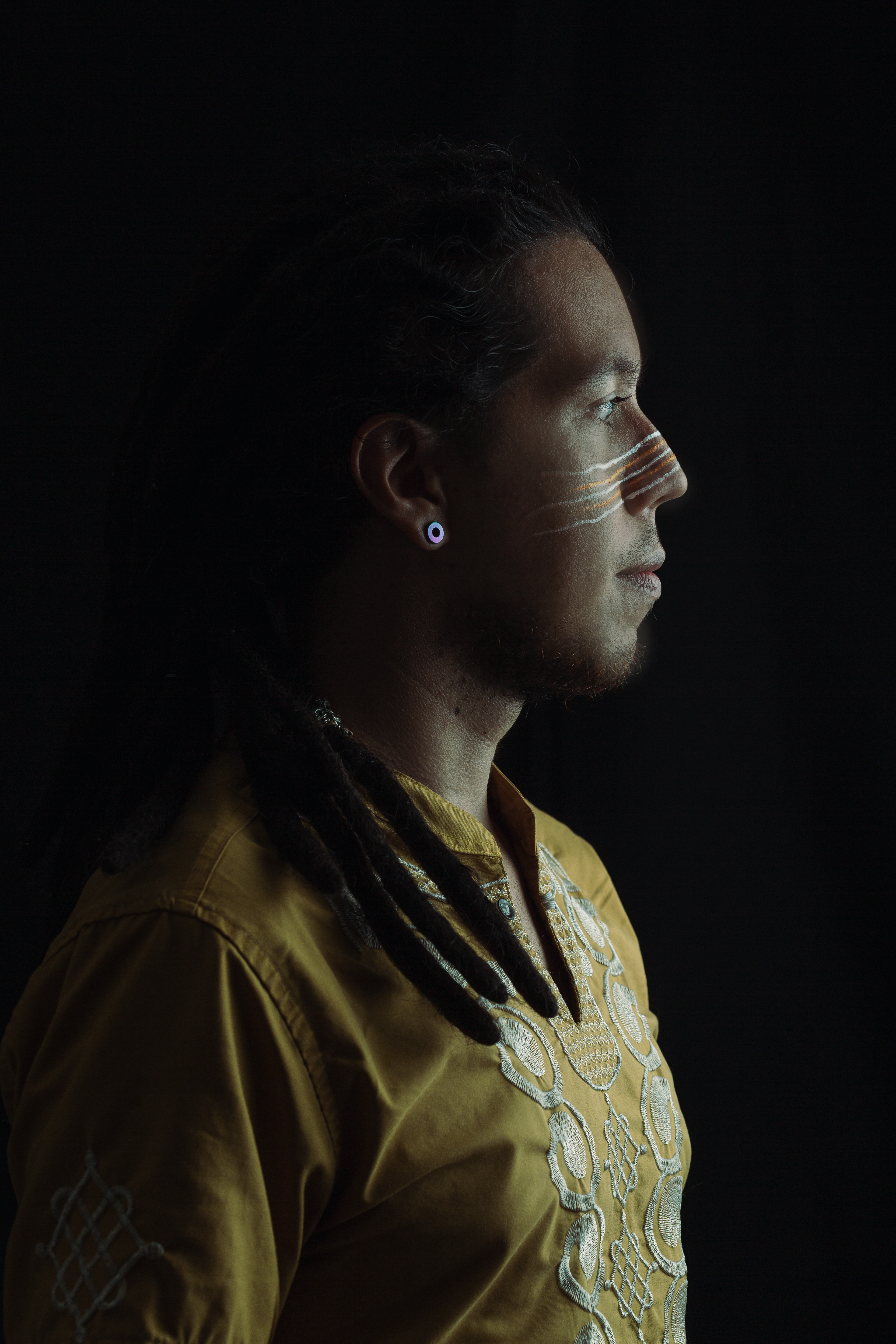 Side profile of a man with dreadlocks and facial paint wearing a traditional shirt.