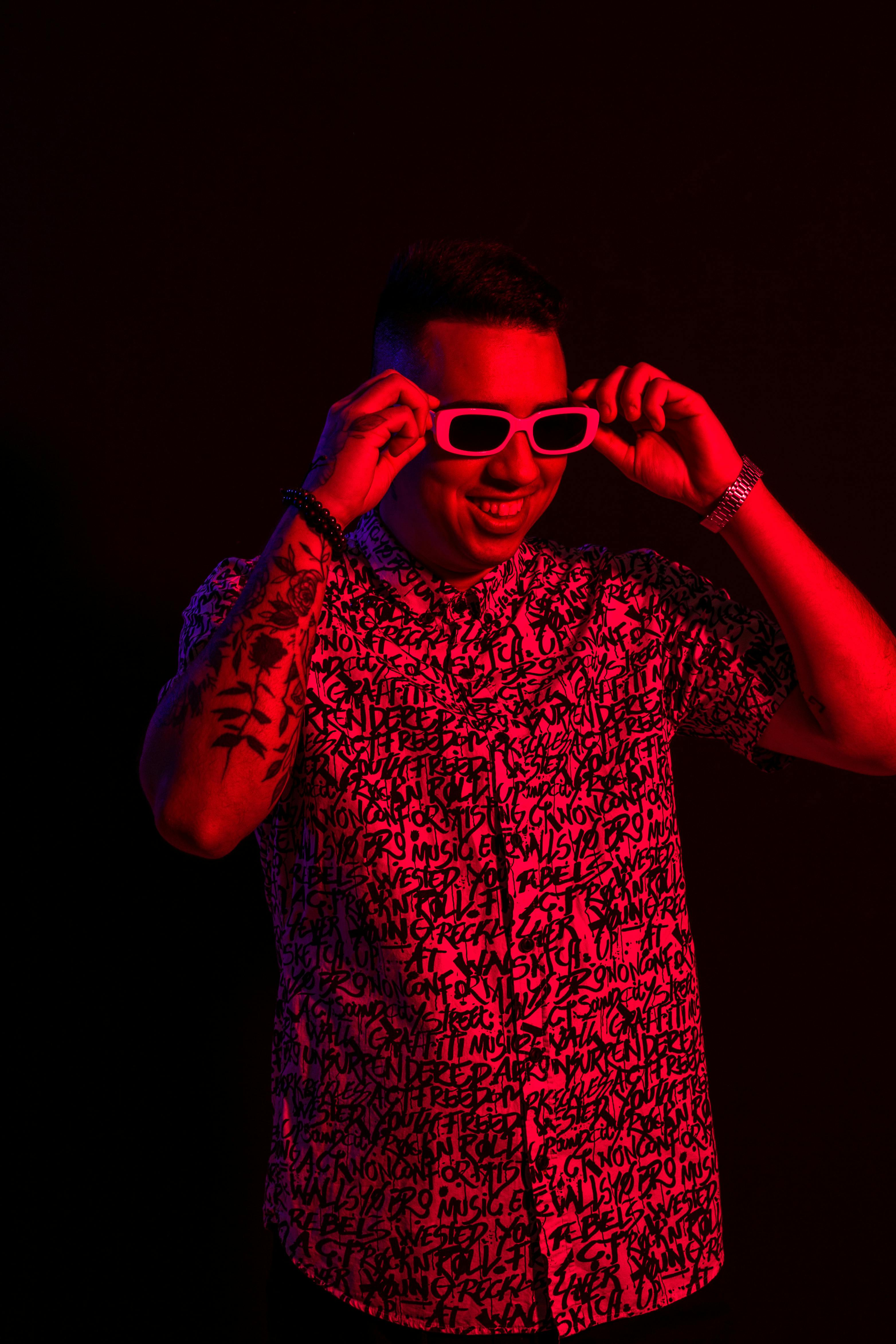 Portrait of a tattooed man smiling in neon red lighting, wearing sunglasses.