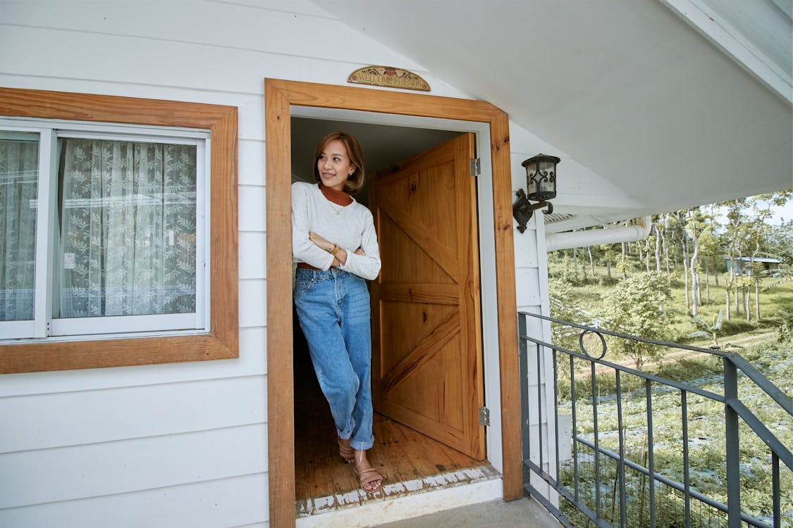 Free Woman in White Long Sleeve Shirt and Blue Denim Jeans Leaning on Brown Wooden Door Stock Photo