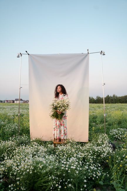 A woman in a floral dress stands in a field holding flowers, framed by a fabric backdrop.