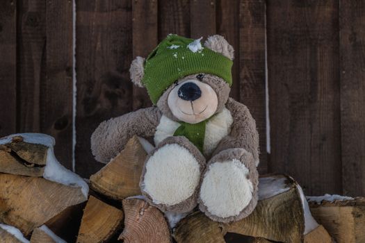 Adorable teddy bear in winter hat sitting on firewood with snow on a rustic wooden background.