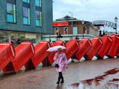 An Elderly Woman in Pink Coat Holding an Umbrella while Walking on the Street Near the Telephone Boxes