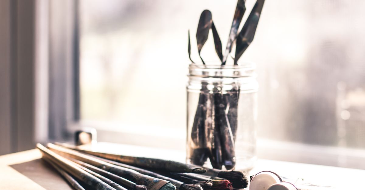 Glass jar with palette knives and paintbrushes on a sunny table, evoking creativity.