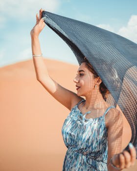 Elegant woman in a blue dress holding a scarf in a sunny desert landscape.