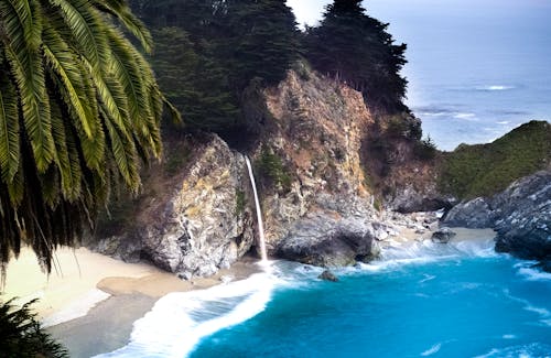 免费 棕色和灰色岩石与绿叶树附近的蓝色大海 素材图片