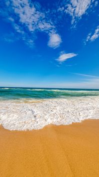 Peaceful beach scene with waves crashing onto the sandy shore under a bright blue sky.