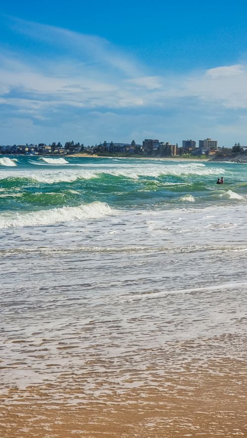 Free Vibrant seaside view with crashing waves, shoreline, and a distant cityscape under a blue sky. Stock Photo