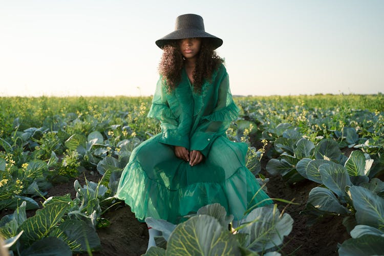 Woman In Green Dress And Black Hat Sitting In Cabbage Field