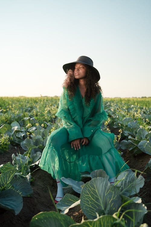 Curly Haired Woman in Hat Sitting on Field of Cabbage