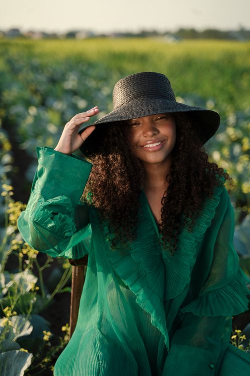 Free A woman in a green dress smiles while sitting in a cabbage field, wearing a wide-brim hat. Stock Photo