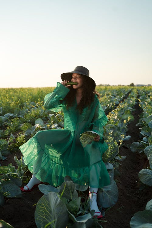 Woman in Green Dress Sitting in Cabbage Field Eating Cucumber