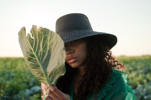 Free Stylish portrait of a woman in a green dress and hat holding a cabbage leaf in a field. Stock Photo