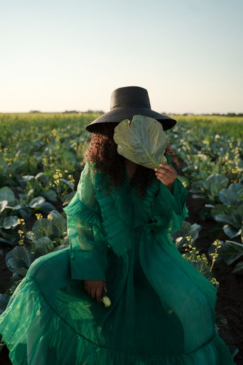 Woman Sitting in Field Hiding Face Behind Cabbage Leaf