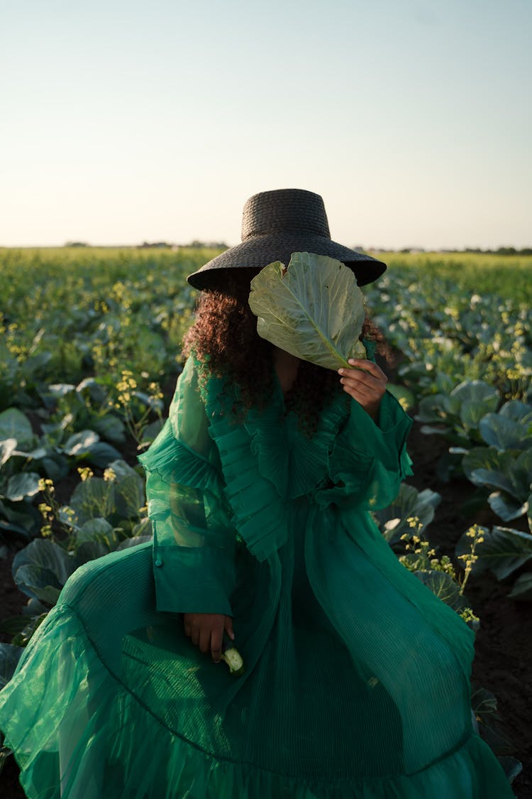 Woman Sitting In Field Hiding Face Behind Cabbage Leaf