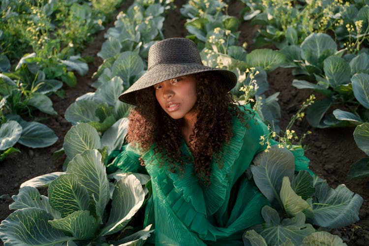 Woman Wearing Green Dress And Brown Hat Sitting Among Rows Of Cabbages In Field