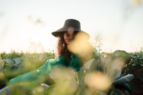 Free Woman in a hat sitting among green plants in a sunlit field, creating a serene outdoor portrait. Stock Photo