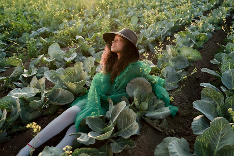 Young Woman In Green Dress And Brown Hat Sitting With Legs Stretched In Cabbage Field