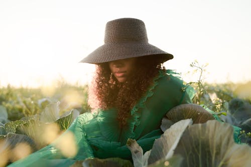 Woman in Green Dress Sitting in Cabbage Field with Brown Hat Covering Her Eyes