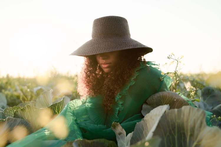 Woman In Green Dress Sitting In Cabbage Field With Brown Hat Covering Her Eyes