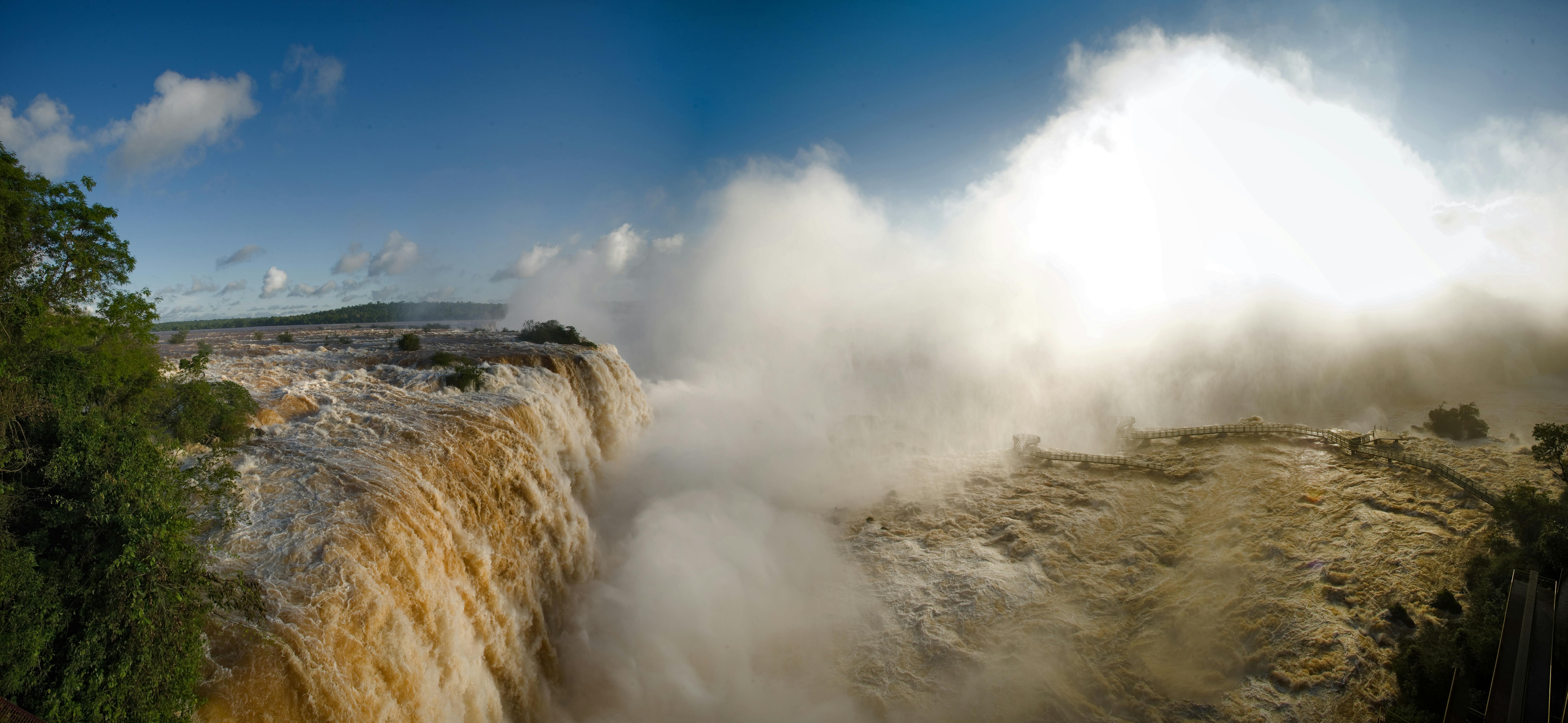 Panoramic view of the powerful Iguazu Falls surrounded by mist and lush greenery under a clear blue sky.