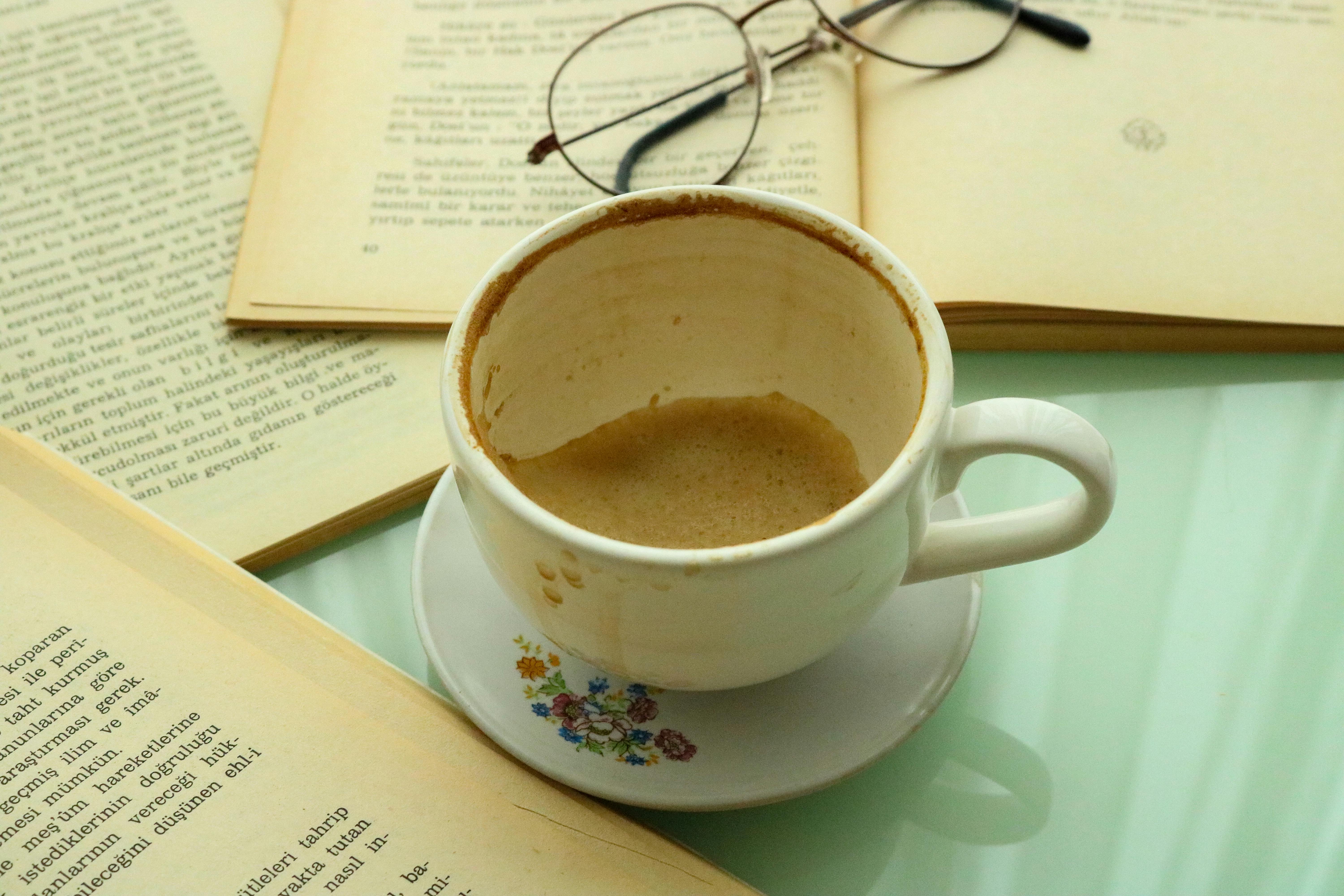 An empty coffee cup surrounded by open books and glasses on a glass table.