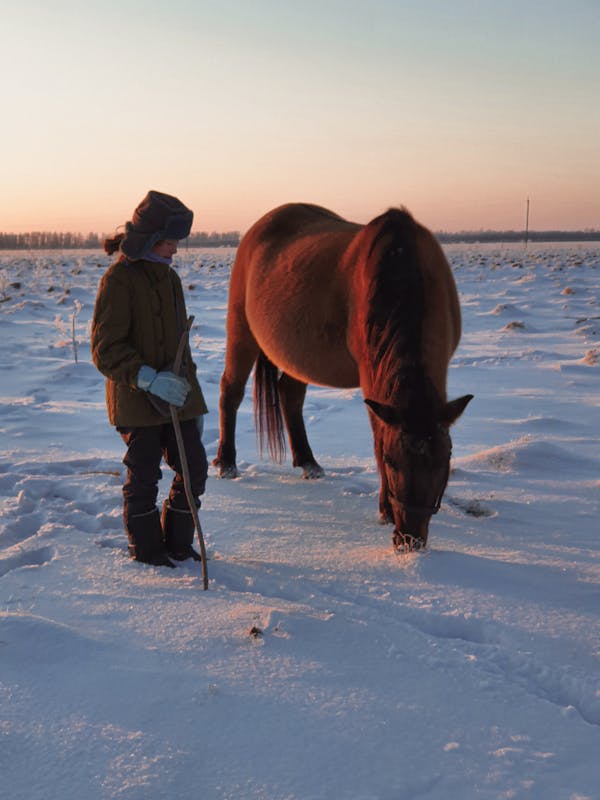 Unrecognizable Person in Warm Clothes Standing by Horse in Snowy Field