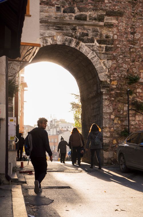 Free A bustling urban street scene with people walking under a historic stone archway at sunset. Stock Photo