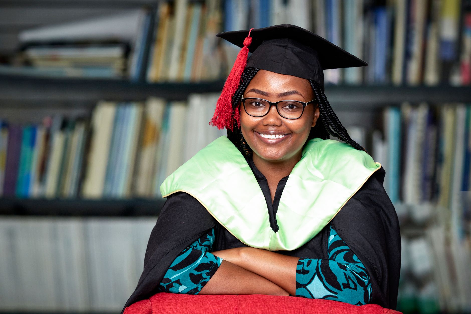 Smiling woman in graduation gown and cap sitting in a library, looking confident.